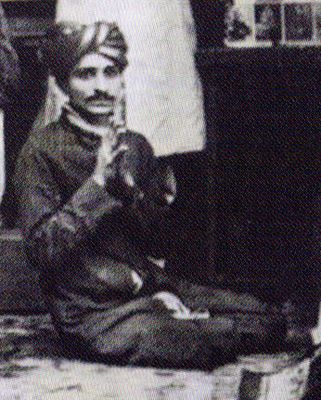 Seated cross-legged in turban and dark coat.
